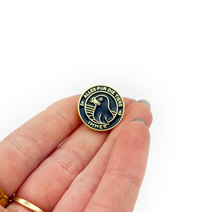 Person hält einen "Alles für die Tiere" Ansteck-Pin aus Emaille in Blau und Gold mit "Alles für die Tiere, immer" Schriftaufzug, sowie Robben-Motiv im der Hand.