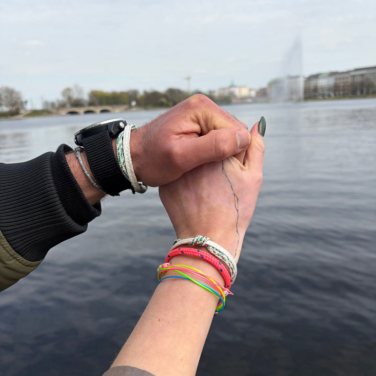 Zwei Personen halten sich an der Hand und tragen beide das Enard Bay Bracenet aus Fischernetz in Weiß und Grün mit Umwicklung in der Mitte und mit Bajonettverschluss in Silber am Handgelenk. 