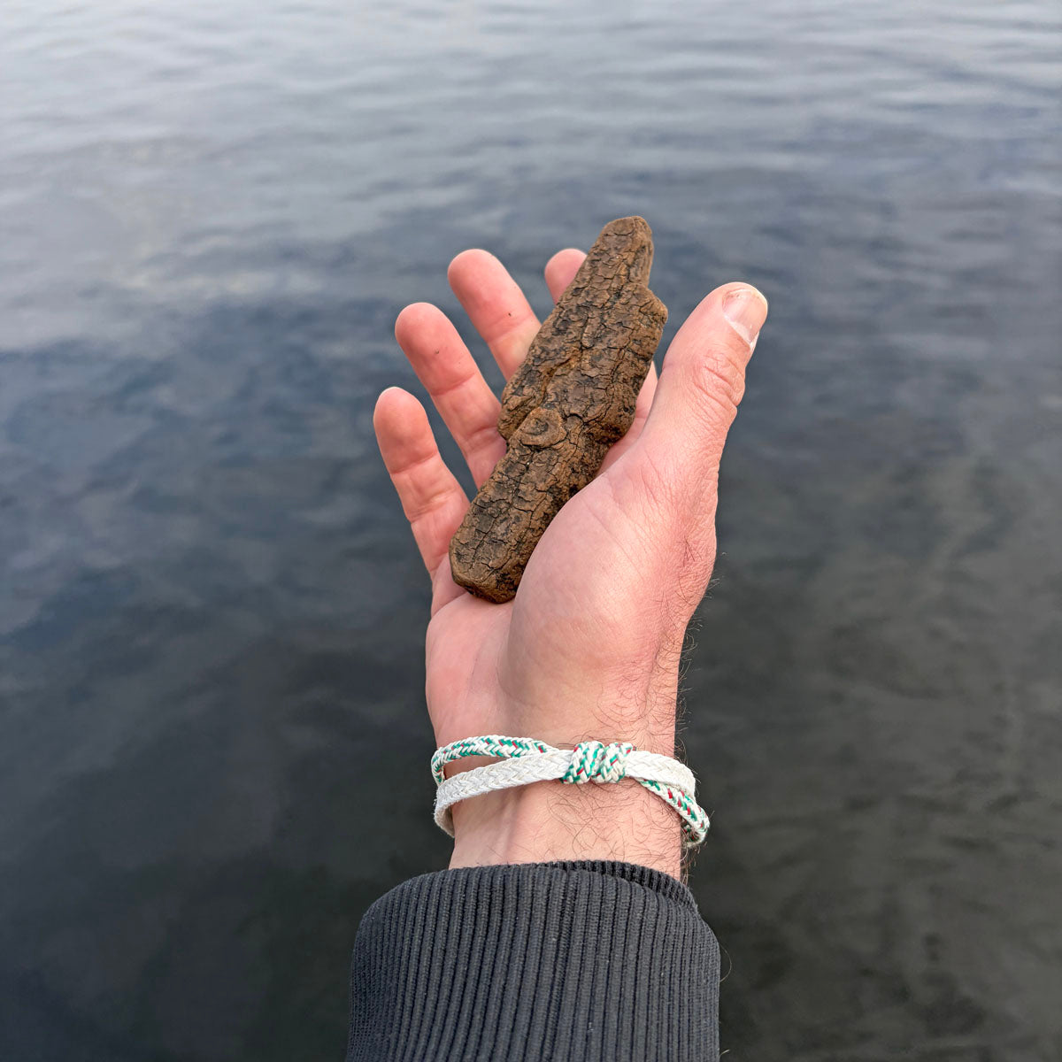Person trägt das Enard Bay Bracenet aus Fischernetz in Weiß und Grün mit Umwicklung in der Mitte und mit Bajonettverschluss in Silber und hat ein Stück Holz in der Hand. 