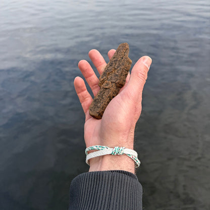 Person trägt das Enard Bay Bracenet aus Fischernetz in Weiß und Grün mit Umwicklung in der Mitte und mit Bajonettverschluss in Silber und hat ein Stück Holz in der Hand. 