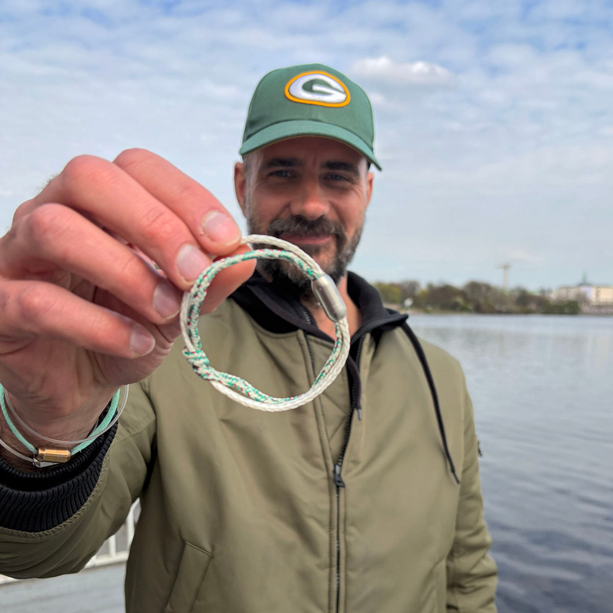 Person hält das Enard Bay Bracenet aus Fischernetz in Weiß und Grün mit Umwicklung in der Mitte und mit Bajonettverschluss in Silber vor das Gesicht und lächelt. 