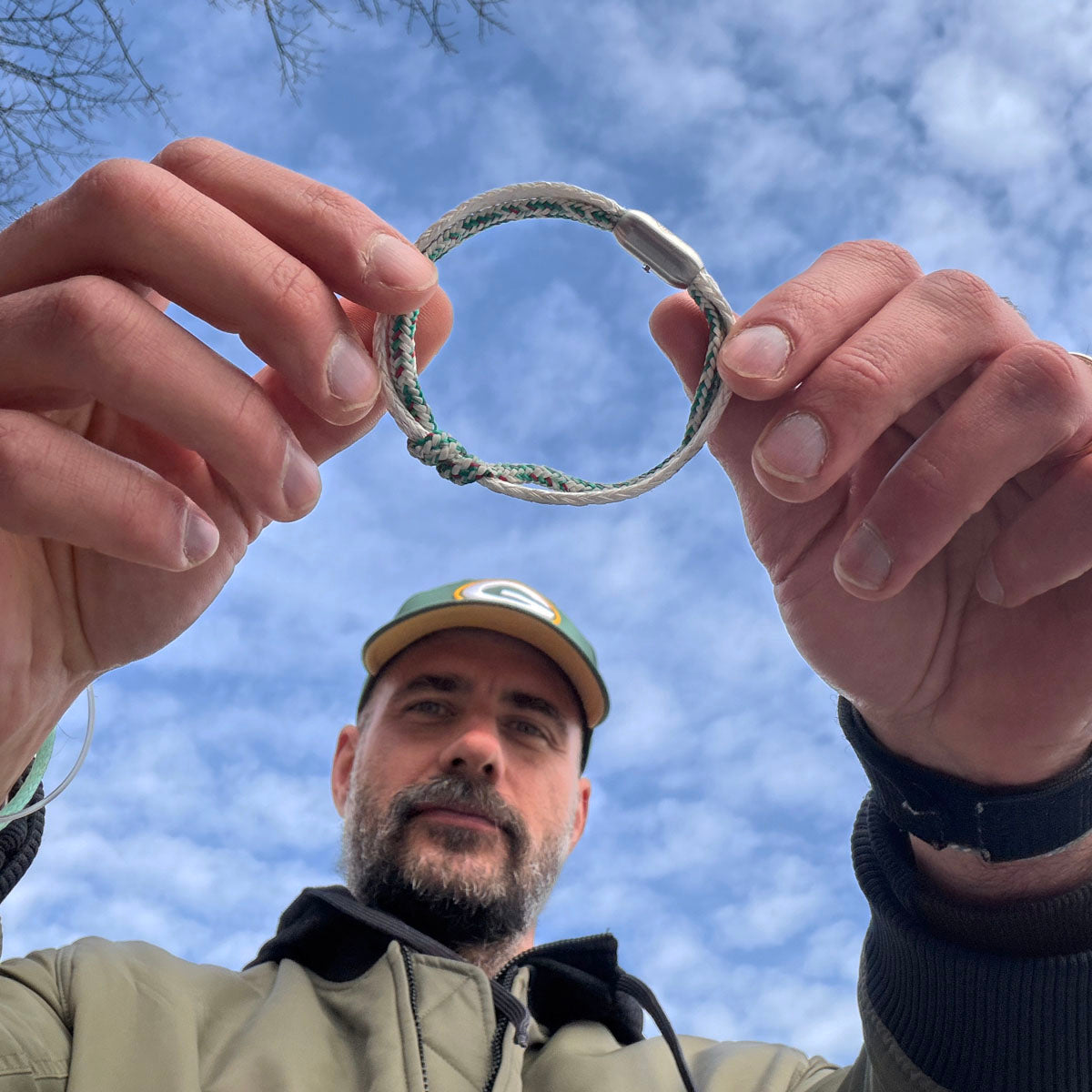 Person hält das Enard Bay Bracenet aus Fischernetz in Weiß und Grün mit Umwicklung in der Mitte und mit Bajonettverschluss in Silber in den Händen und schaut in die Kamera. 