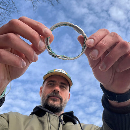 Person hält das Enard Bay Bracenet aus Fischernetz in Weiß und Grün mit Umwicklung in der Mitte und mit Bajonettverschluss in Silber in den Händen und schaut in die Kamera. 