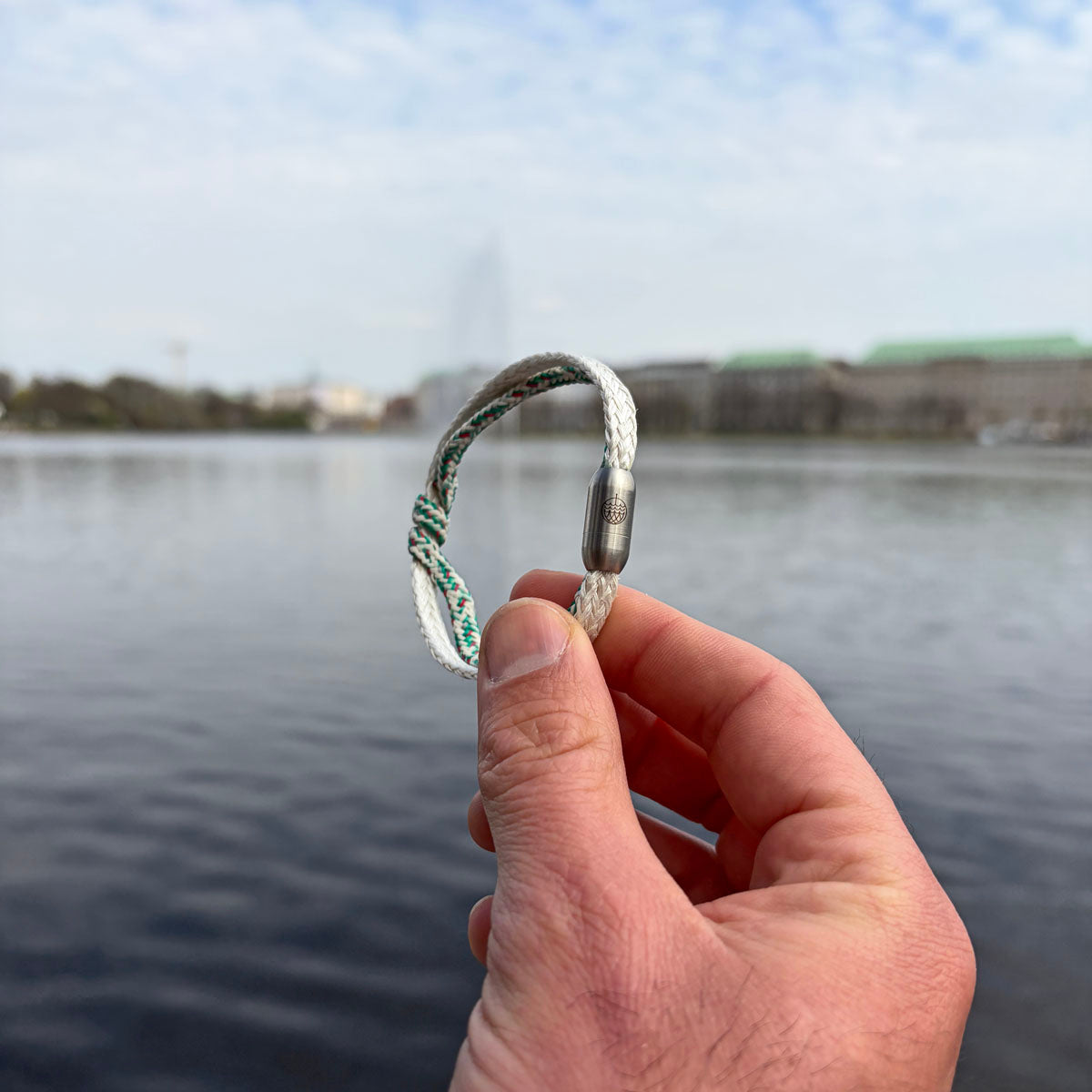Person hält das Enard Bay Bracenet aus Fischernetz in Weiß und Grün mit Umwicklung in der Mitte und mit Bajonettverschluss in Silber in der Hand. Im Hintergrund sieht man Wasser. 