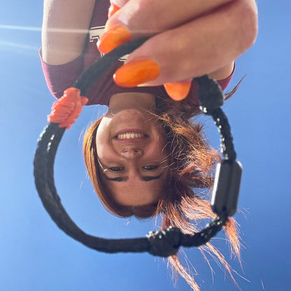 Person hält das BRACENET Upcycling Armband Pumpkin Bay II aus schwarzen alten Fischernetzen mit einem Knoten aus orangenen Dolly Ropes und einem schwarzen Magnetverschluss vors Gesicht und lacht. Im Hintergrund ist der blaue Himmel.