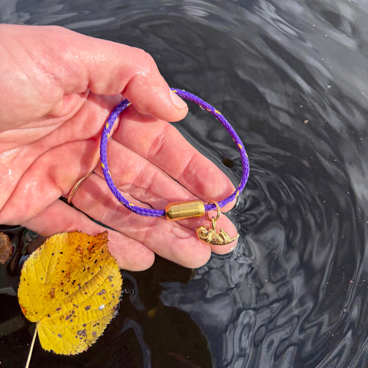 Person hält das BRACENET Armband aus Fischernetz in Lila mit Magnetverschluss in und einem Otter-Anhänger in Gold. in der Hand über dem Wasser.