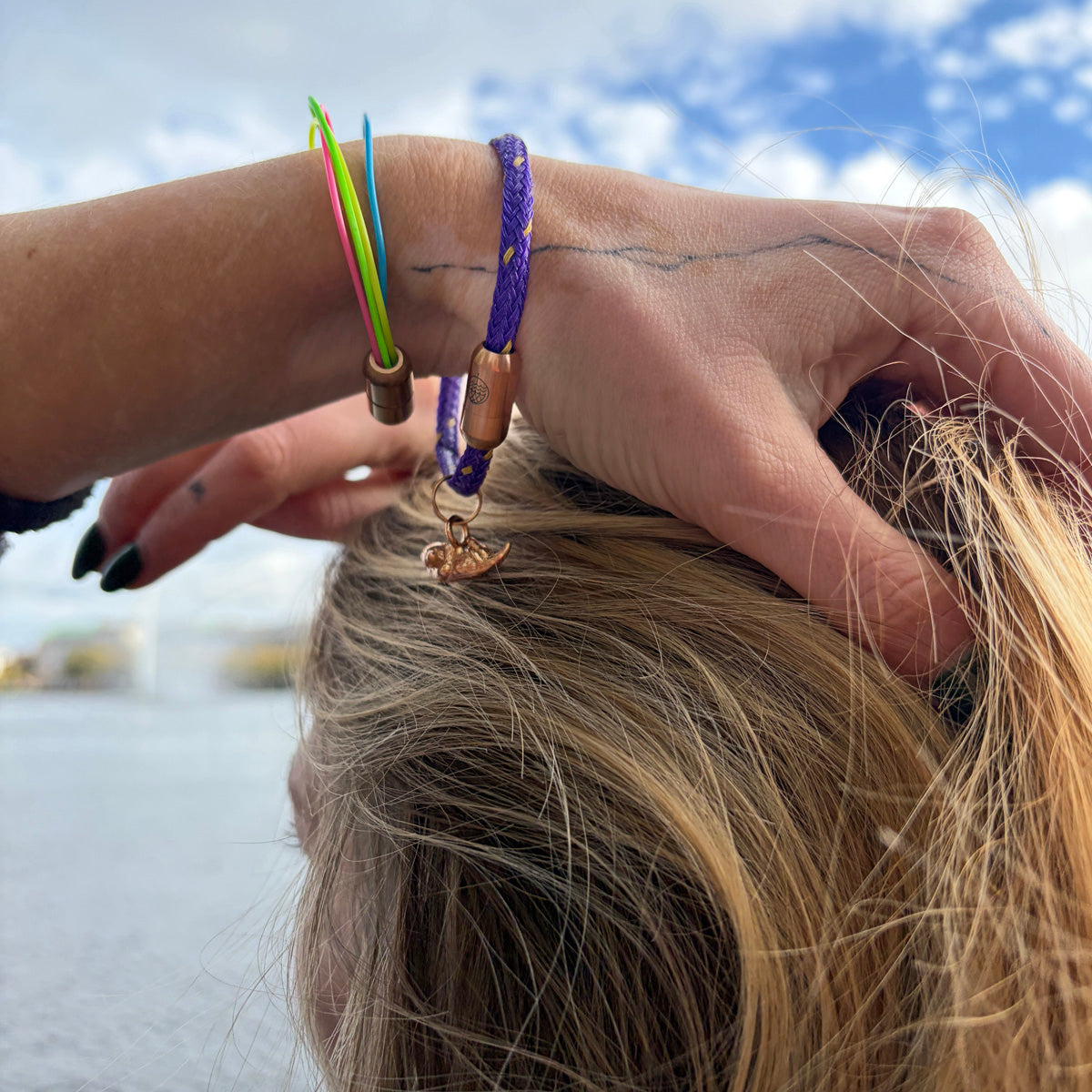 Person trägt das BRACENET Armband aus Fischernetz in Lila mit Magnetverschluss in Roségold und einem Otter-Anhänger in Roségold am Handgelenk und fasst sich in die Haare.