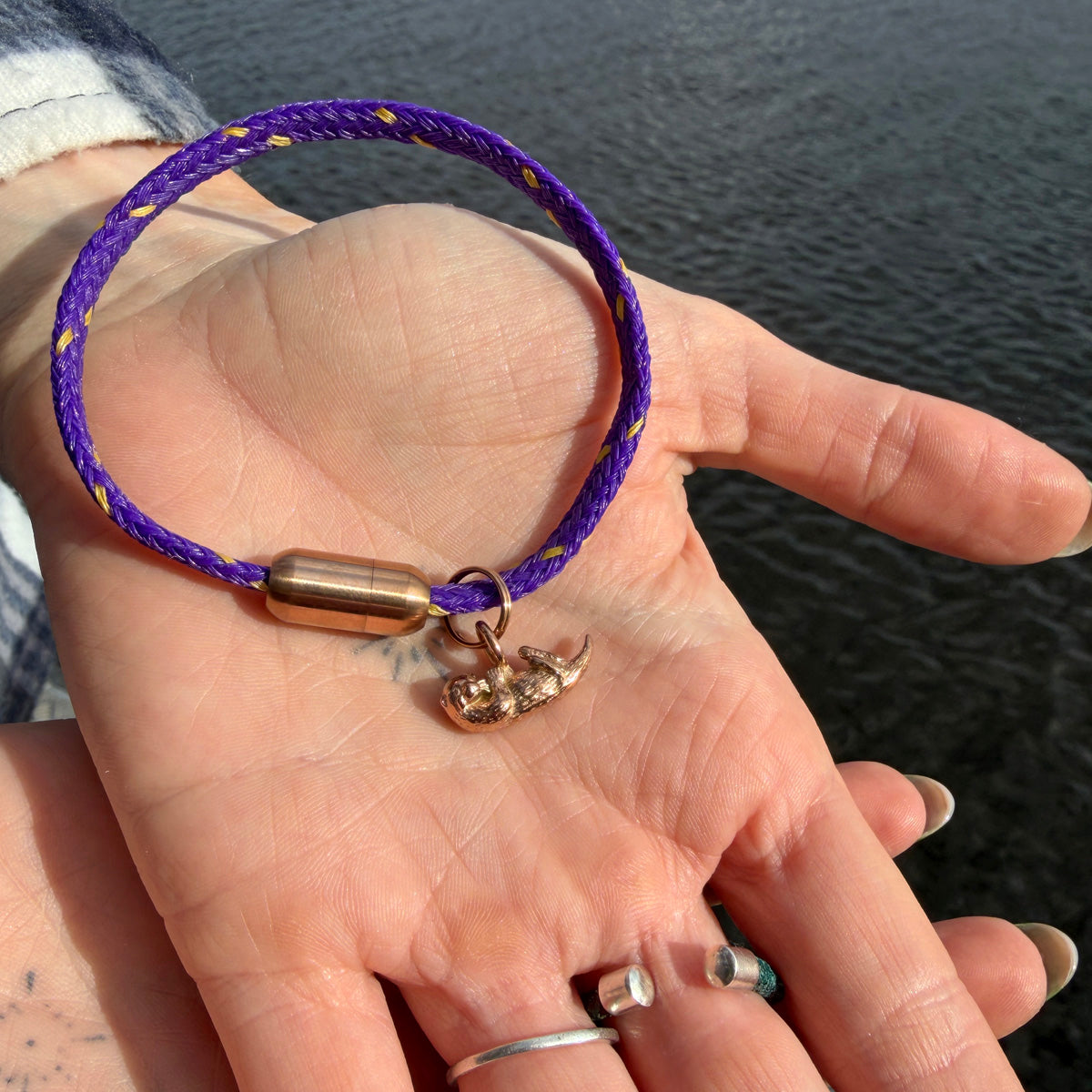 Person hält das BRACENET Armband aus Fischernetz in Lila mit Magnetverschluss in Roségold und einem Otter-Anhänger in Roségold in der Handfläche über dem Wasser. 