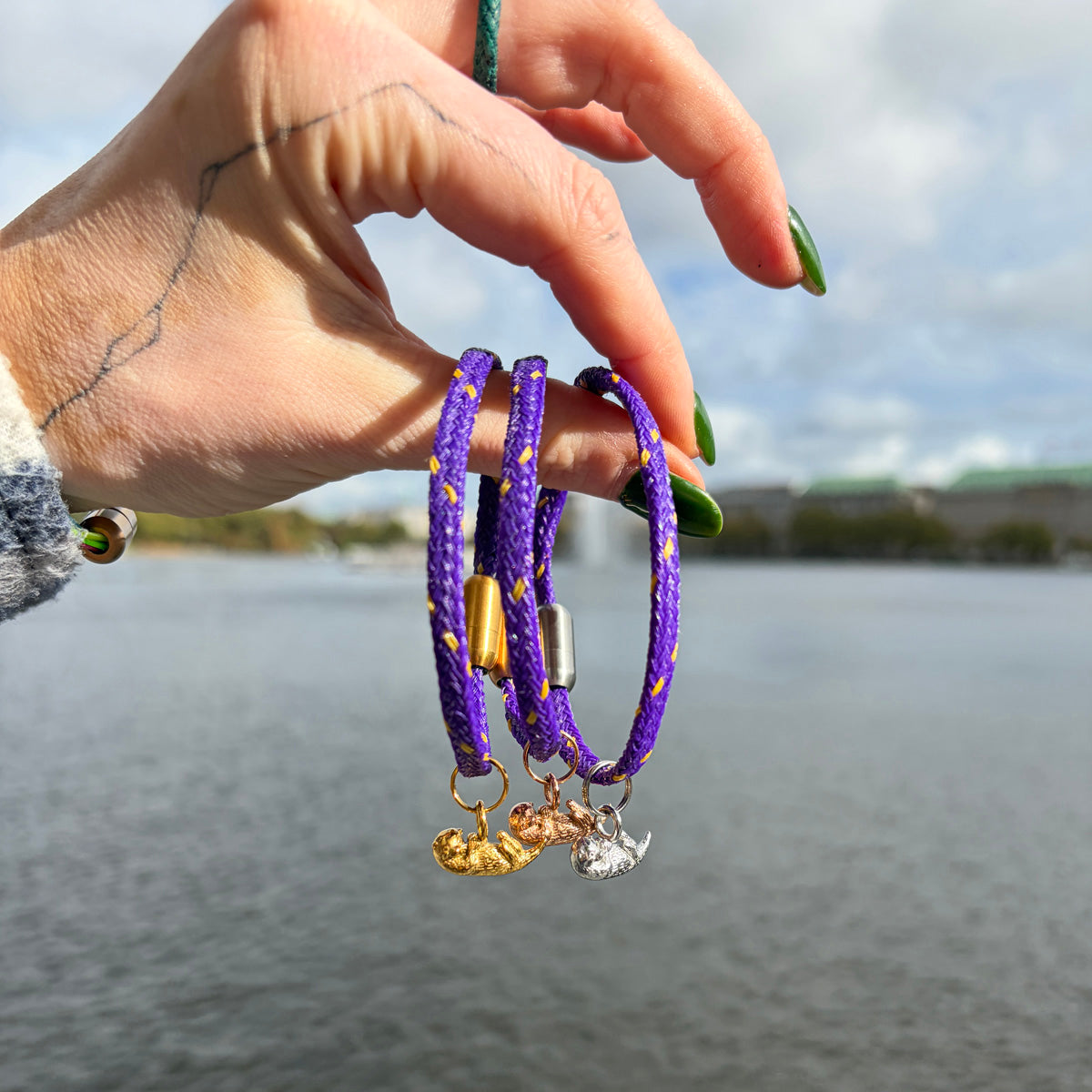 Person hält drei BRACENET Armband aus Fischernetz in Lila mit Magnetverschluss in Silber, Roségold oder Gold und einem Otter-Anhänger in Silber, Roségold und Gold zwischen den Fingern, über dem Wasser.