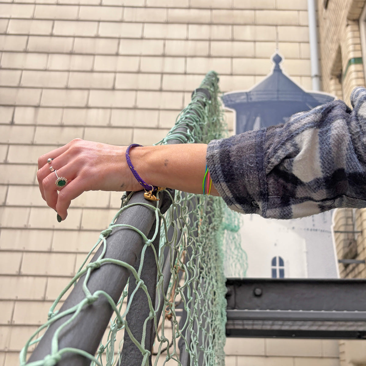 Person hat die Hand auf ein Geländer gelegt und trägt das BRACENET Armband aus Fischernetz in Lila mit Magnetverschluss in Gold und einem Otter-Anhänger in Gold am Handgelenk. 