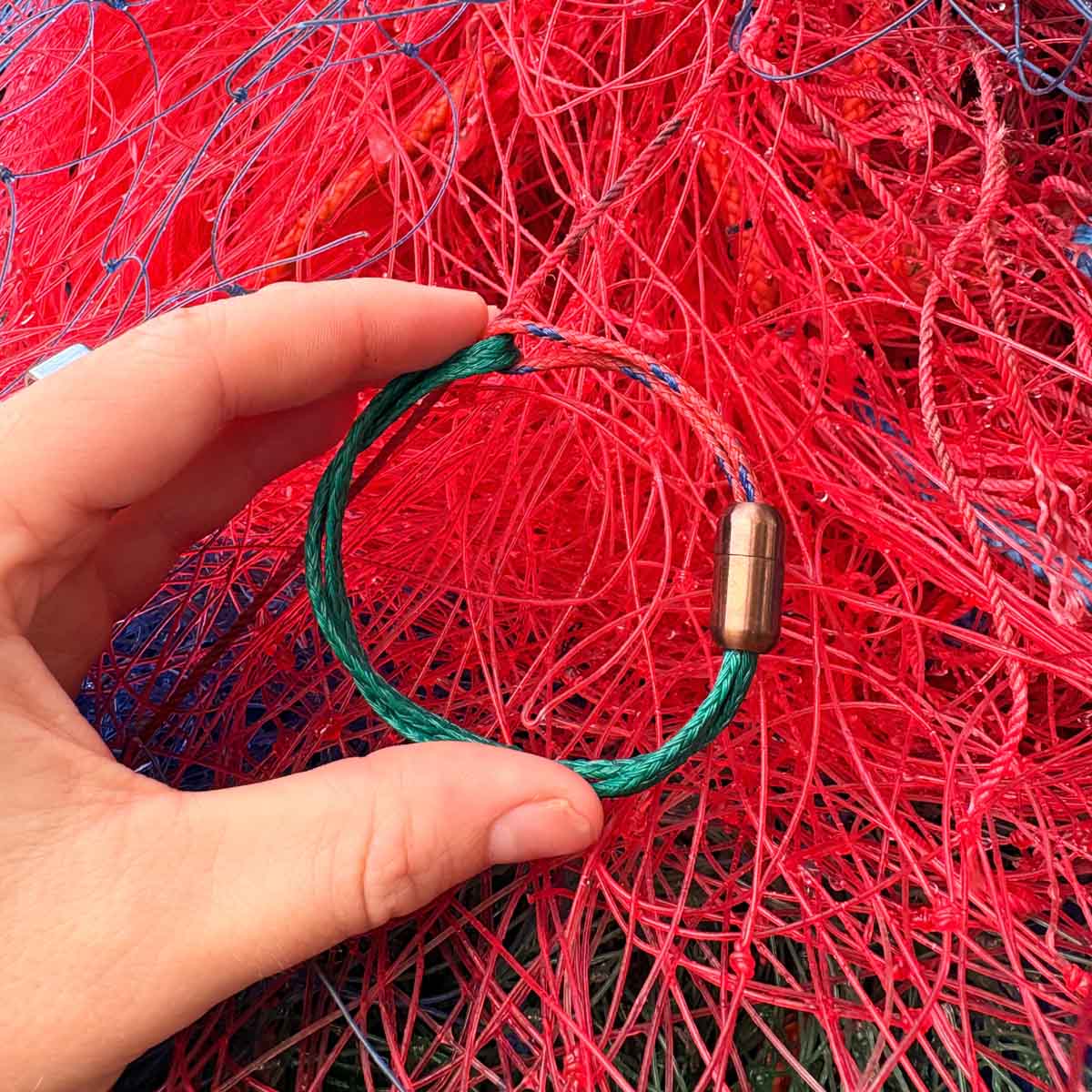 Person hält das BRACENET Christmas Bracenet aus einem grünen und einem roten Fischernetz mit coffee Magnetverschluss vor rotes Fischernetz.