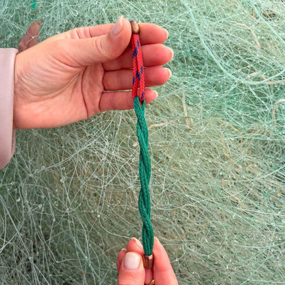 Person hält das BRACENET Christmas Bracenet aus einem grünen und einem roten Fischernetz mit coffee Magnetverschluss gestreckt vor Fischernetzen. Man sieht, dass das grüne Netz gedreht ist. 