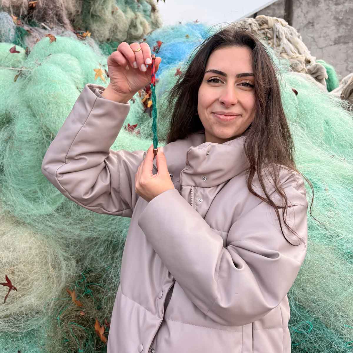 Person hält das BRACENET Christmas Bracenet aus einem grünen und einem roten Fischernetz mit coffee Magnetverschluss gestreckt neben ihr Gesicht. Im Hintergrund sind Fischernetze.