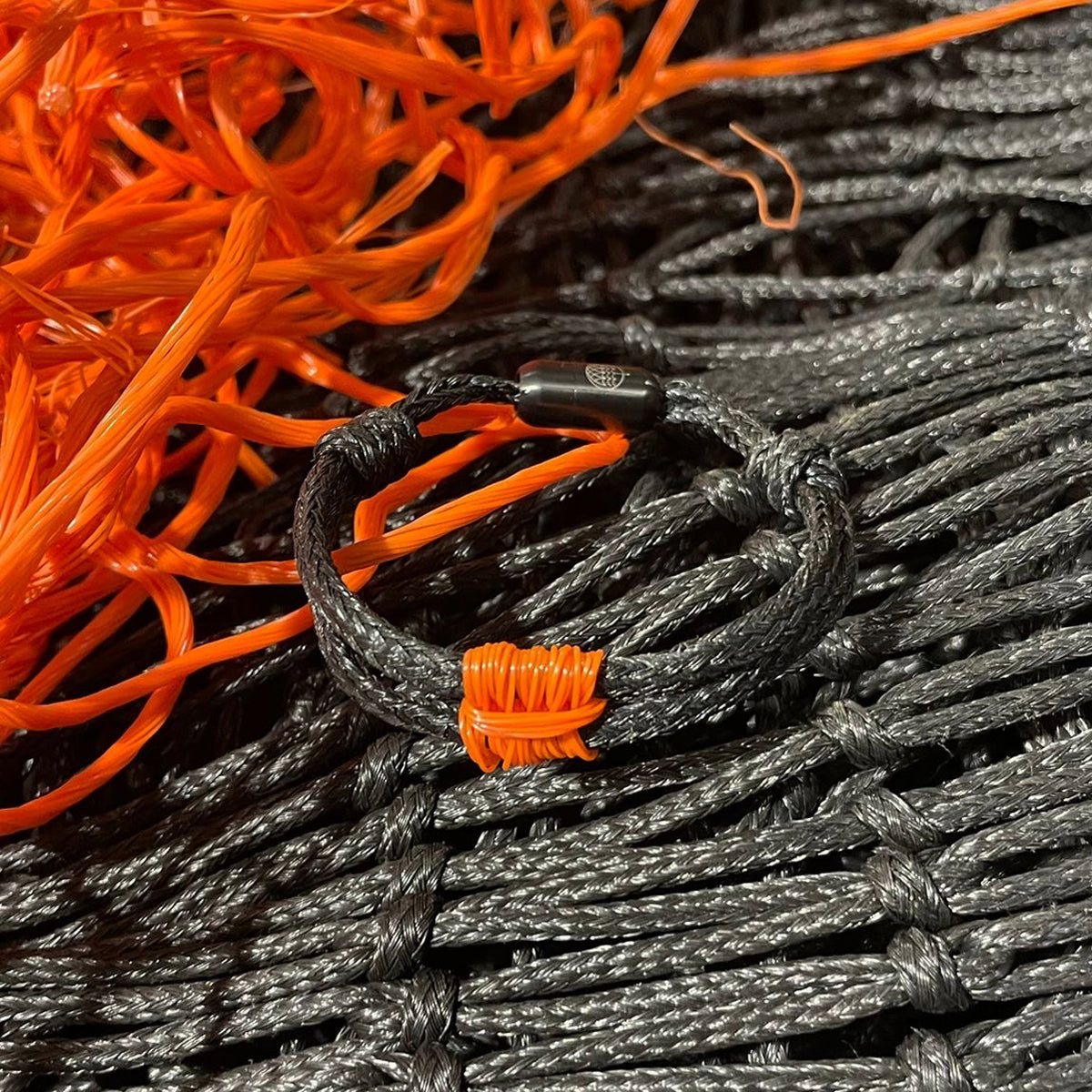 BRACENET Upcycling Armband Pumpkin Bay II aus schwarzen alten Fischernetzen mit einem Knoten aus orangenen Dolly Ropes und einem schwarzen Magnetverschluss liegt auf dem schwarzen und orangenen Fischernetz. 
