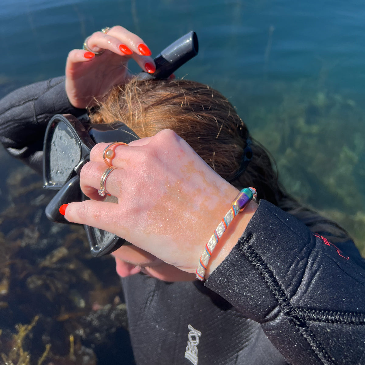 Person im Neoprenanzug kommt aus dem Wasser und trägt das BRACENET Upcycling Armband Rainbow Bay aus alten weißen Fischernetzen mit bunten Dolly Ropes umwickelt und bunten Magnetverschluss mit Farbverlauf am Handgelenk