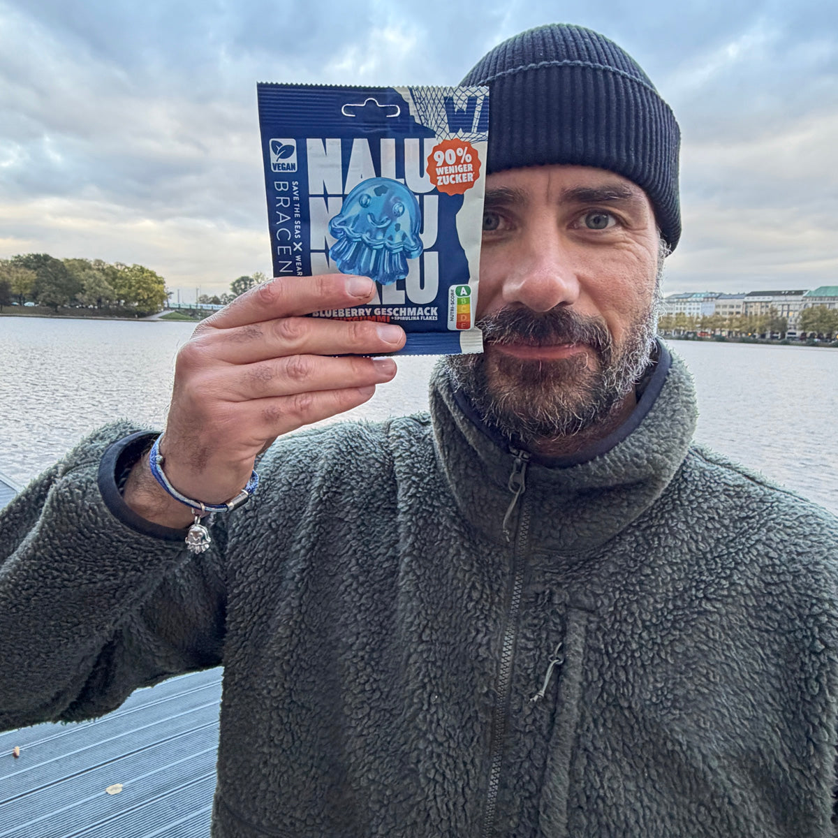 Eine Person Benjamin hält eine Fruchtgummi Tüte von Nalu in der Hand. Am Handgelenk trägt er ein Nalu Bracenet Armband mit Quallen Anhänger in silber. Im Hintergrund ist die Alster zu sehen.