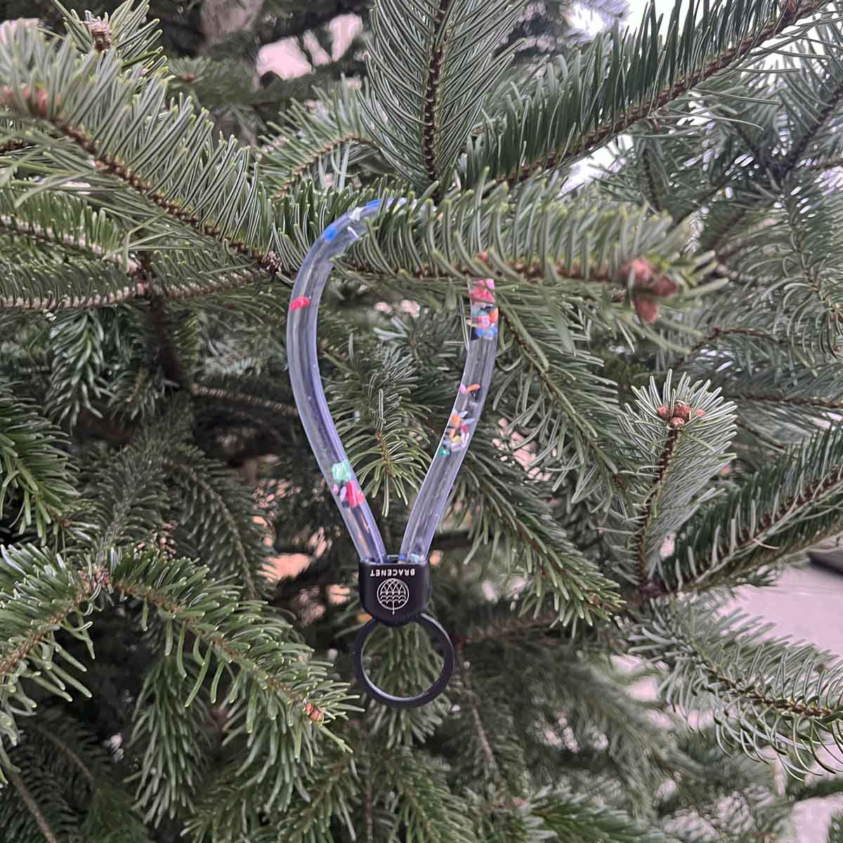BRACENET Floating Upcycling Schlüsselanhänger aus alten Plastiktuben gefüllt mit Mikroplastik, destilliertem Wasser und Glycerin mit Edelstahlkappe und Ring in schwarz hängt an einem Tannenbaum.