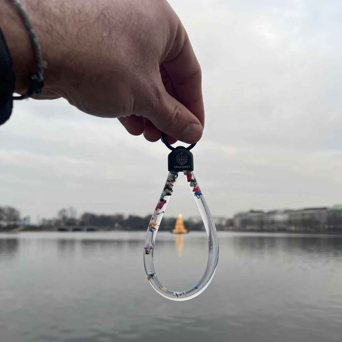 Person hält BRACENET Floating Upcycling Schlüsselanhänger aus alten Plastiktuben gefüllt mit Mikroplastik, destilliertem Wasser und Glycerin mit Edelstahlkappe und Ring in schwarz am Ring fest. Durch den Schlüsselanhänger hindurch sieht man einen beleuchteten Weihnachtsbaum.