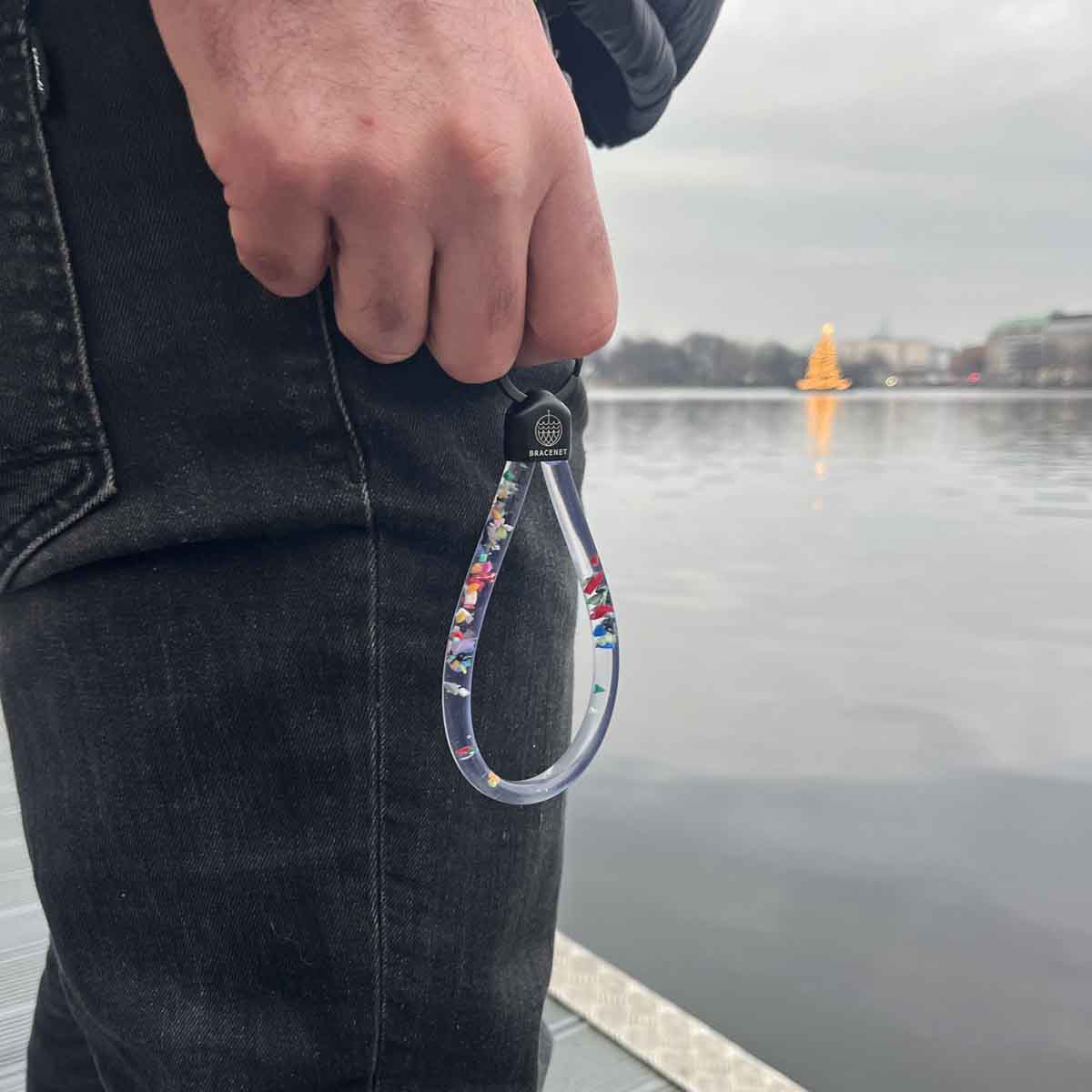Person hat an einem Finger den BRACENET Floating Upcycling Schlüsselanhänger aus alten Plastiktuben gefüllt mit Mikroplastik, destilliertem Wasser und Glycerin mit Edelstahlkappe und Ring in schwarz hängen. 