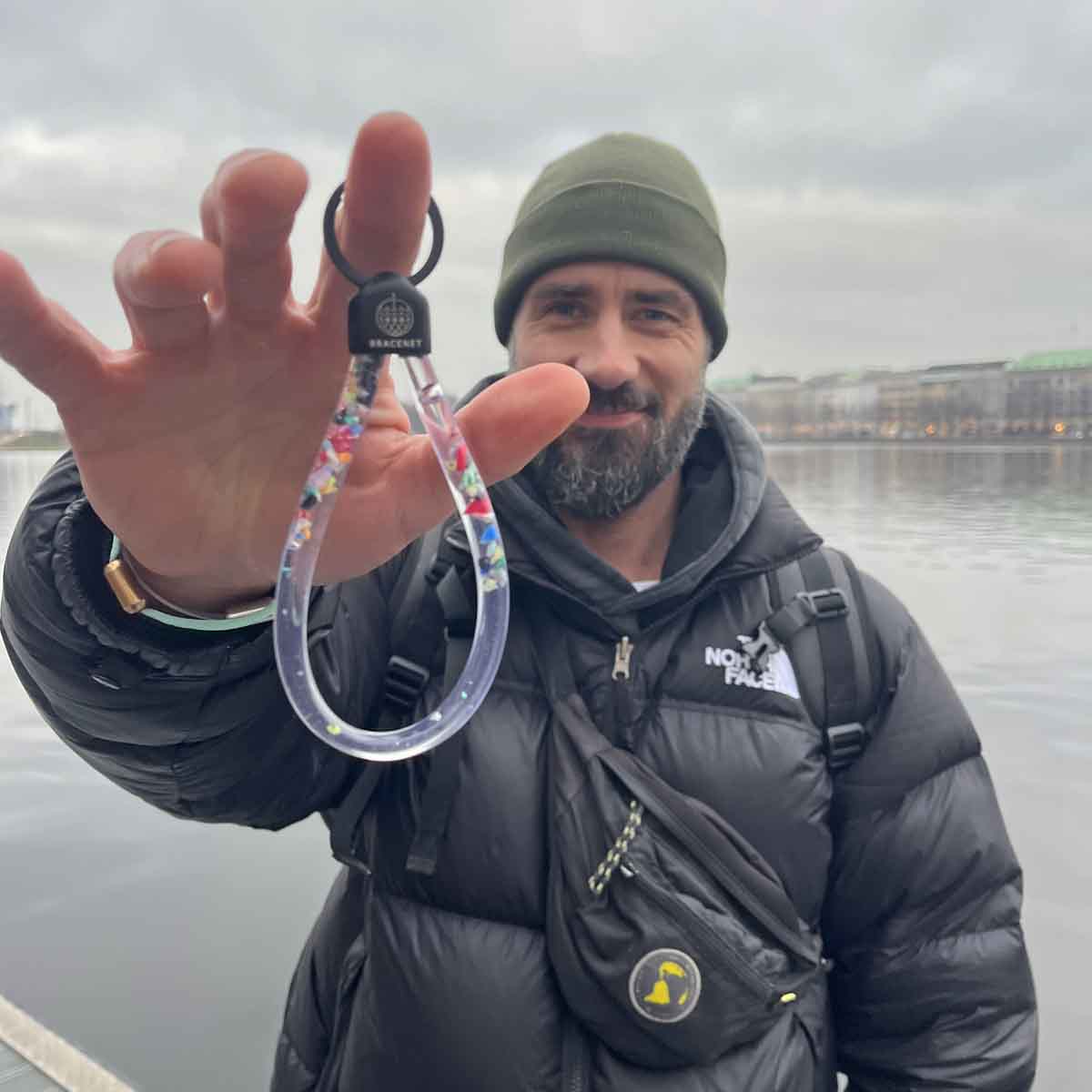 Person hält den BRACENET Floating Upcycling Schlüsselanhänger aus alten Plastiktuben gefüllt mit Mikroplastik, destilliertem Wasser und Glycerin mit Edelstahlkappe und Ring in schwarz vor sich und grinst. 