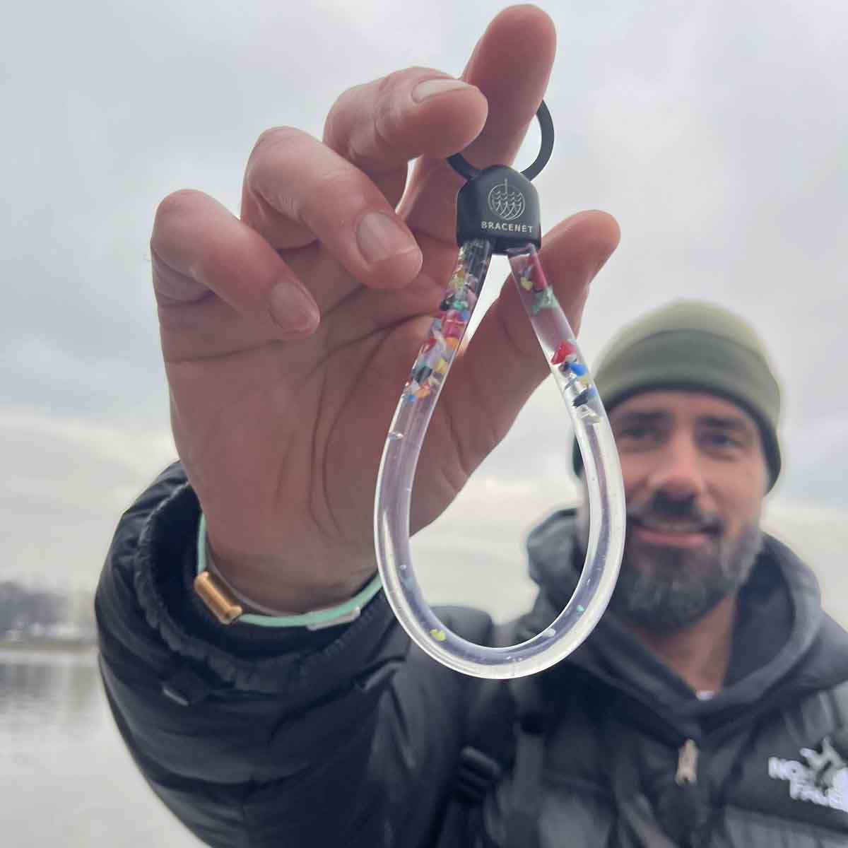 Person hält den BRACENET Floating Upcycling Schlüsselanhänger aus alten Plastiktuben gefüllt mit Mikroplastik, destilliertem Wasser und Glycerin mit Edelstahlkappe und Ring in schwarz an einem Finger vor sich und lacht. 