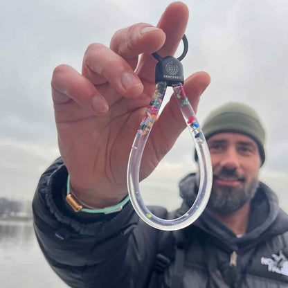 Person hält den BRACENET Floating Upcycling Schlüsselanhänger aus alten Plastiktuben gefüllt mit Mikroplastik, destilliertem Wasser und Glycerin mit Edelstahlkappe und Ring in schwarz an einem Finger vor sich und lacht. 