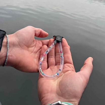 Person hält den BRACENET Floating Upcycling Schlüsselanhänger aus alten Plastiktuben gefüllt mit Mikroplastik, destilliertem Wasser und Glycerin mit Edelstahlkappe und Ring in schwarz in den Händen über dem Wasser.