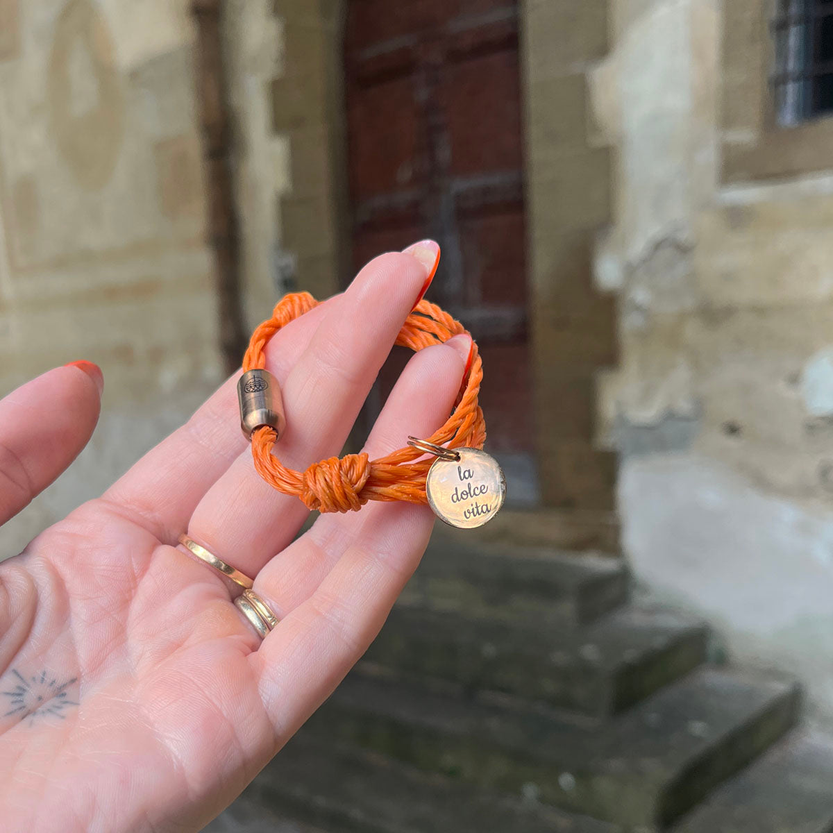 La Dolce Vita Toskana Bracenet Bracelet aus Fischernetz in orange mit Edelstahl Magnetverschluss und einem Edelstahlanhänger mit La Dolce Vita Gravur in der Hand
