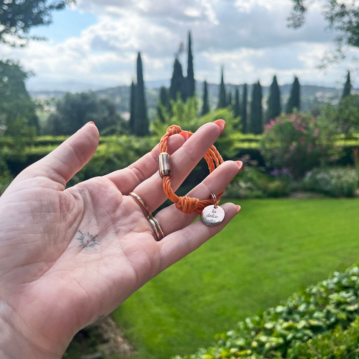 La Dolce Vita Toskana Bracenet Bracelet aus Fischernetz in orange mit Edelstahl Magnetverschluss und einem Edelstahlanhänger mit La Dolce Vita Gravur in der Hand gehalten