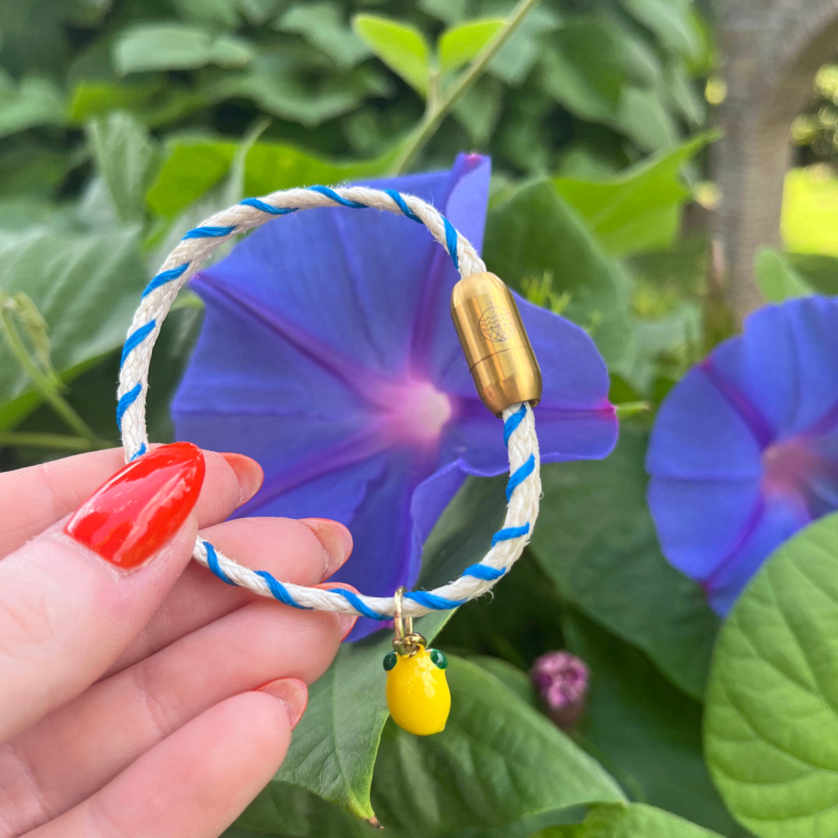 Almalfi Bracenet Bracelet aus Fischernetz in weiß und blau mit Edelstahl Magnetverschluss und einem Zitronenanhänger in der Hand mit Blume im Hintergrund