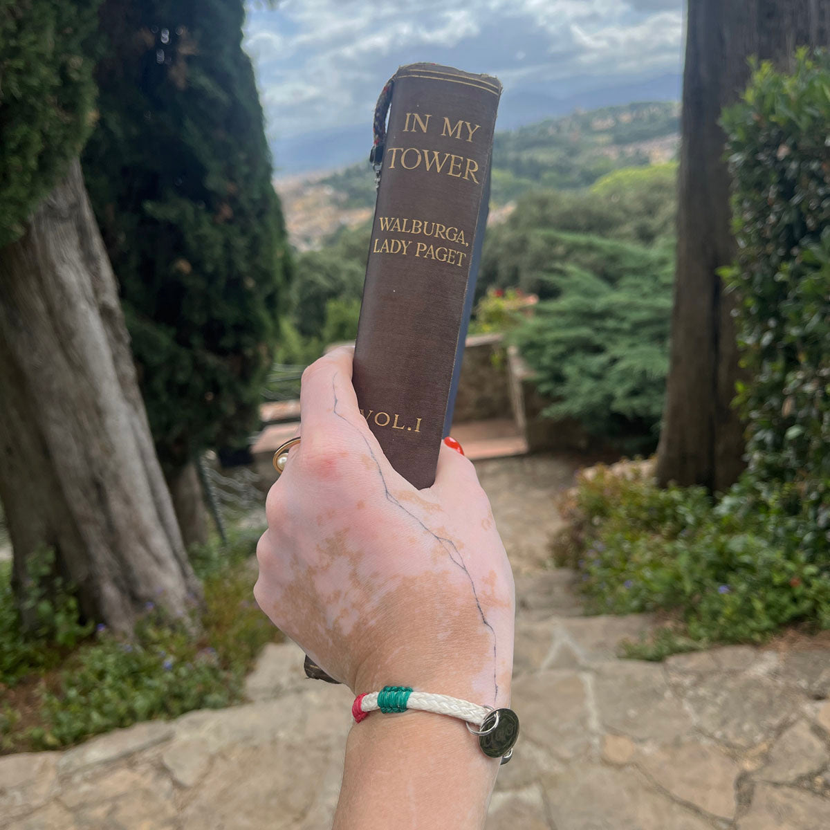 Italy, Italien Bracenet Bracelet  aus ehemaligem Fischernetz in Weiß mit roten und grünen Akzenten mit Edelstahl Magnetverschluss und einem Edelstahlanhänger mit Ciao Gravur am Handgelenk mit Buch in der Hand
