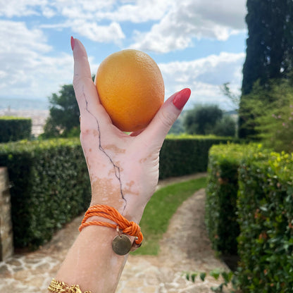La Dolce Vita Toskana Bracenet Bracelet aus Fischernetz in orange mit Edelstahl Magnetverschluss und einem Edelstahlanhänger mit La Dolce Vita Gravur am Handgelenk mit Orange in der Hand