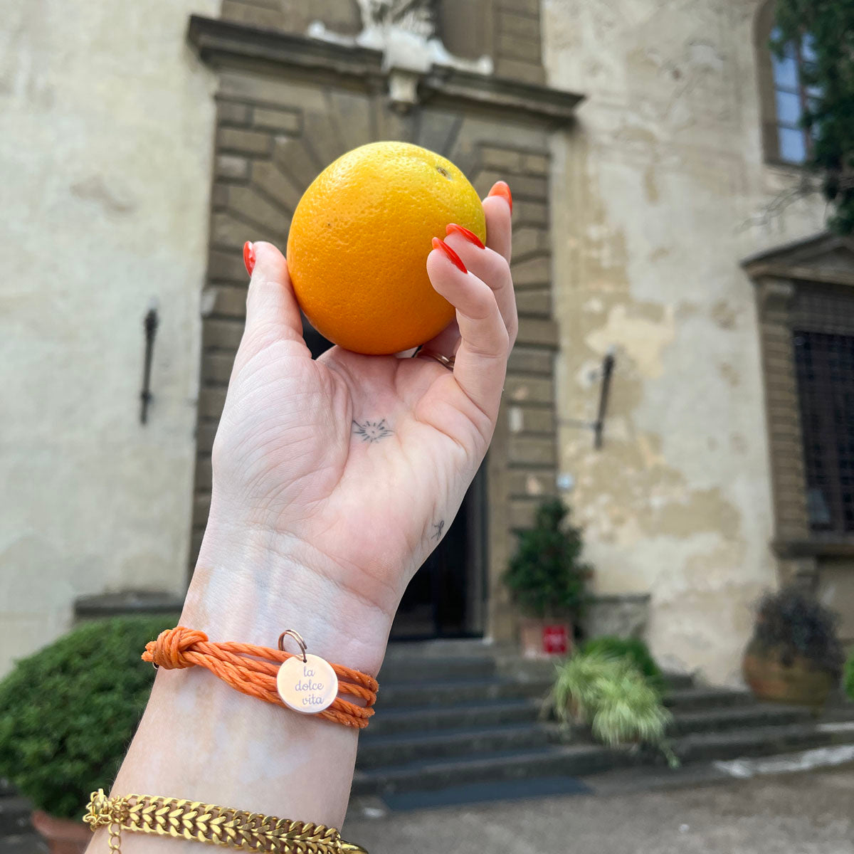 La Dolce Vita Toskana Bracenet Bracelet aus Fischernetz in orange mit Edelstahl Magnetverschluss und einem Edelstahlanhänger mit La Dolce Vita Gravur am Handgelenk und Orange in der Hand