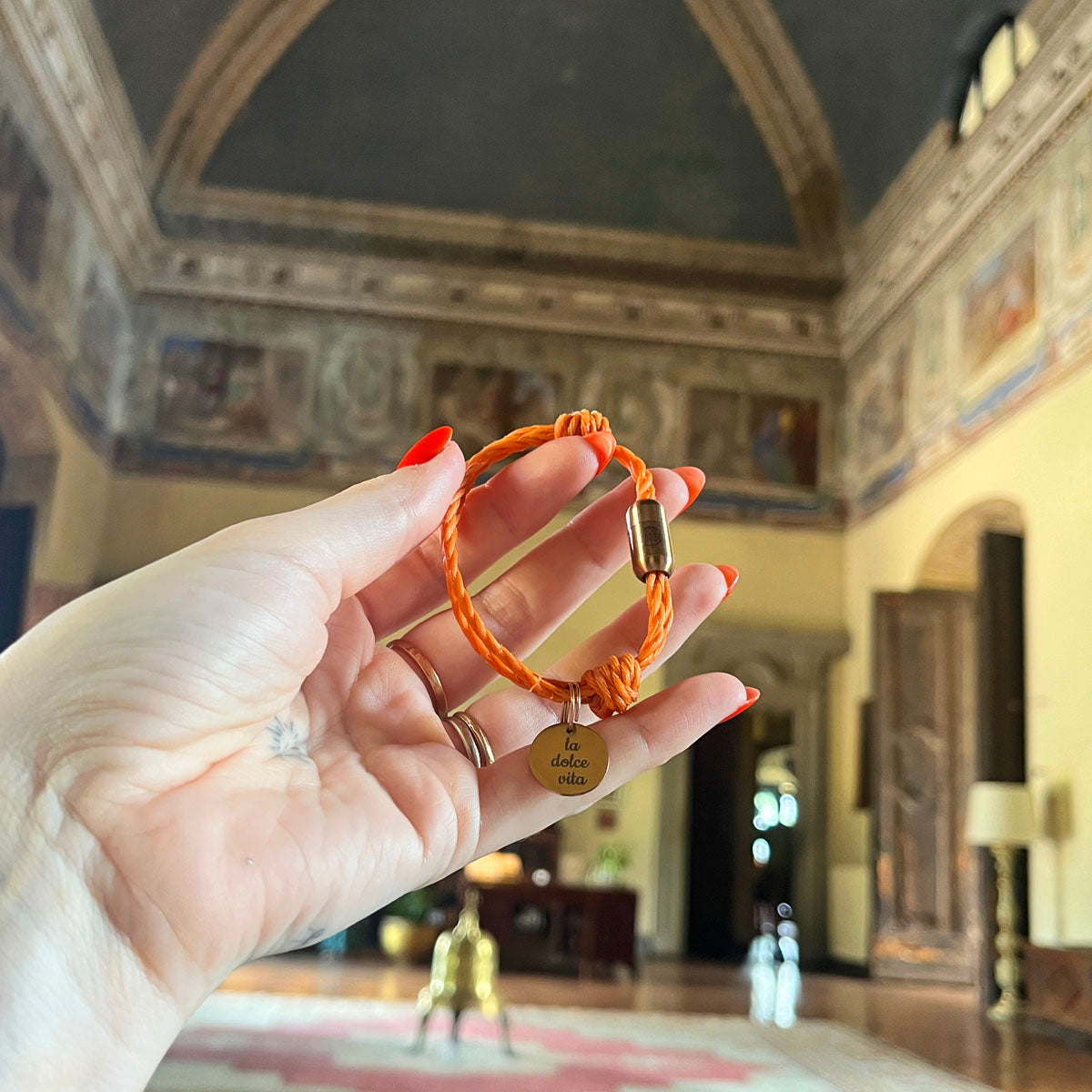 La Dolce Vita Toskana Bracenet Bracelet aus Fischernetz in orange mit Edelstahl Magnetverschluss und einem Edelstahlanhänger mit La Dolce Vita Gravur in der hand gehalten