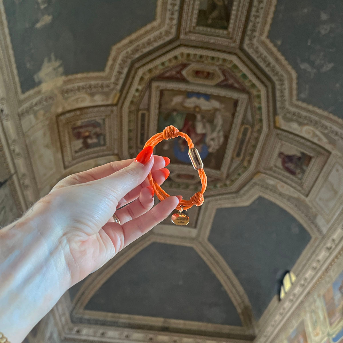 La Dolce Vita Toskana Bracenet Bracelet aus Fischernetz in orange mit Edelstahl Magnetverschluss und einem Edelstahlanhänger mit La Dolce Vita Gravur in der Hand gehalten
