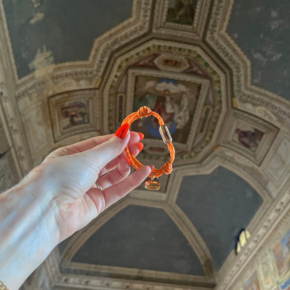 La Dolce Vita Toskana Bracenet Bracelet aus Fischernetz in orange mit Edelstahl Magnetverschluss und einem Edelstahlanhänger mit La Dolce Vita Gravur in der Hand gehalten