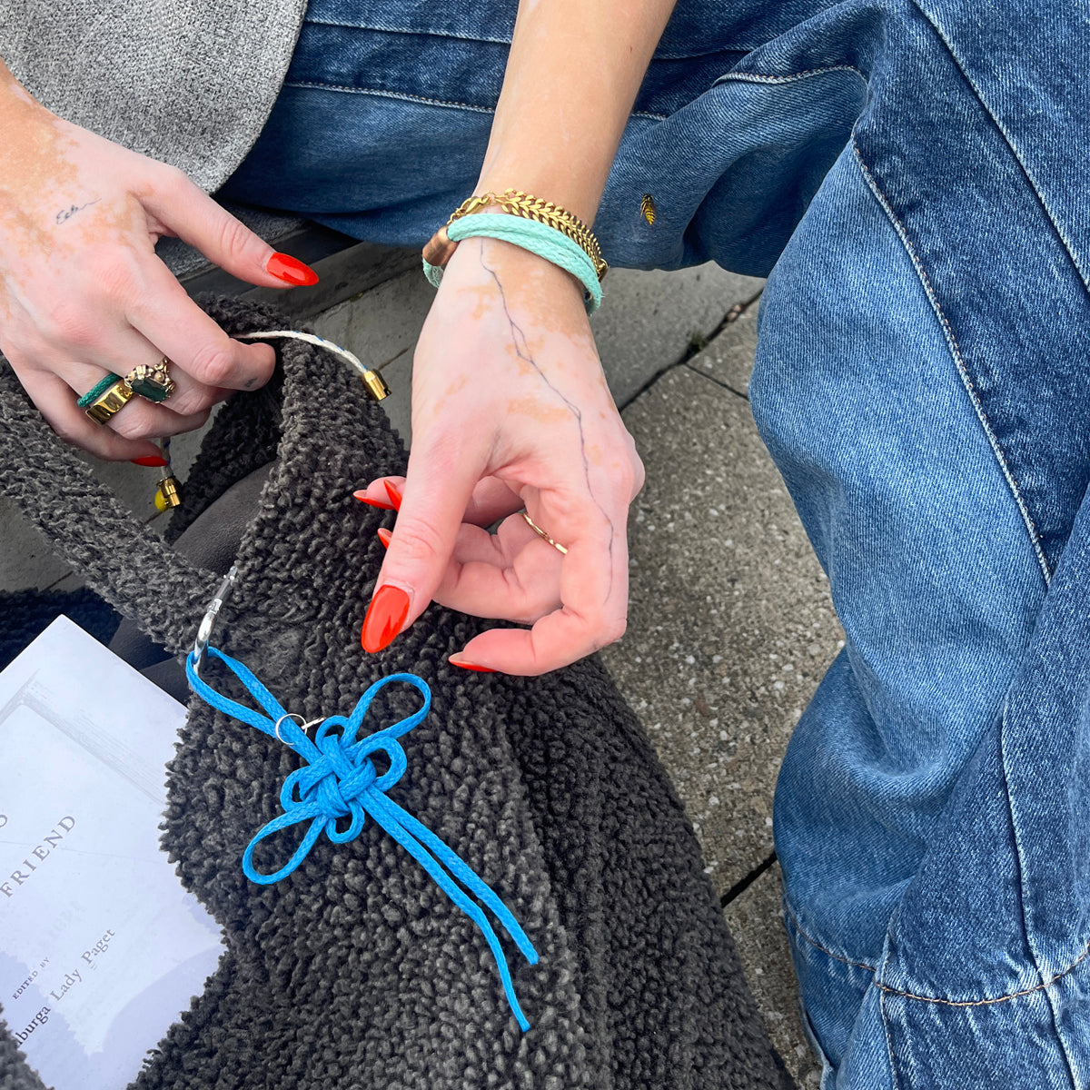 Person befestigt BRACENET Upcycling Taschenanhänger Flores Sea aus blauen alten Fischernetzen mit einem silber Edelstahlanhänger mit BRACENET Logo und einem silber Edelstahlclip in Herzform an einer grauen Tasche. Die Tasche steht auf dem Boden. 