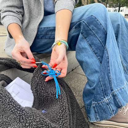 Person sitzt auf einer Treppe und befestigt BRACENET Upcycling Taschenanhänger Flores Sea aus blauen alten Fischernetzen mit einem silber Edelstahlanhänger mit BRACENET Logo und einem silber Edelstahlclip in Herzform an einer grauen Tasche.