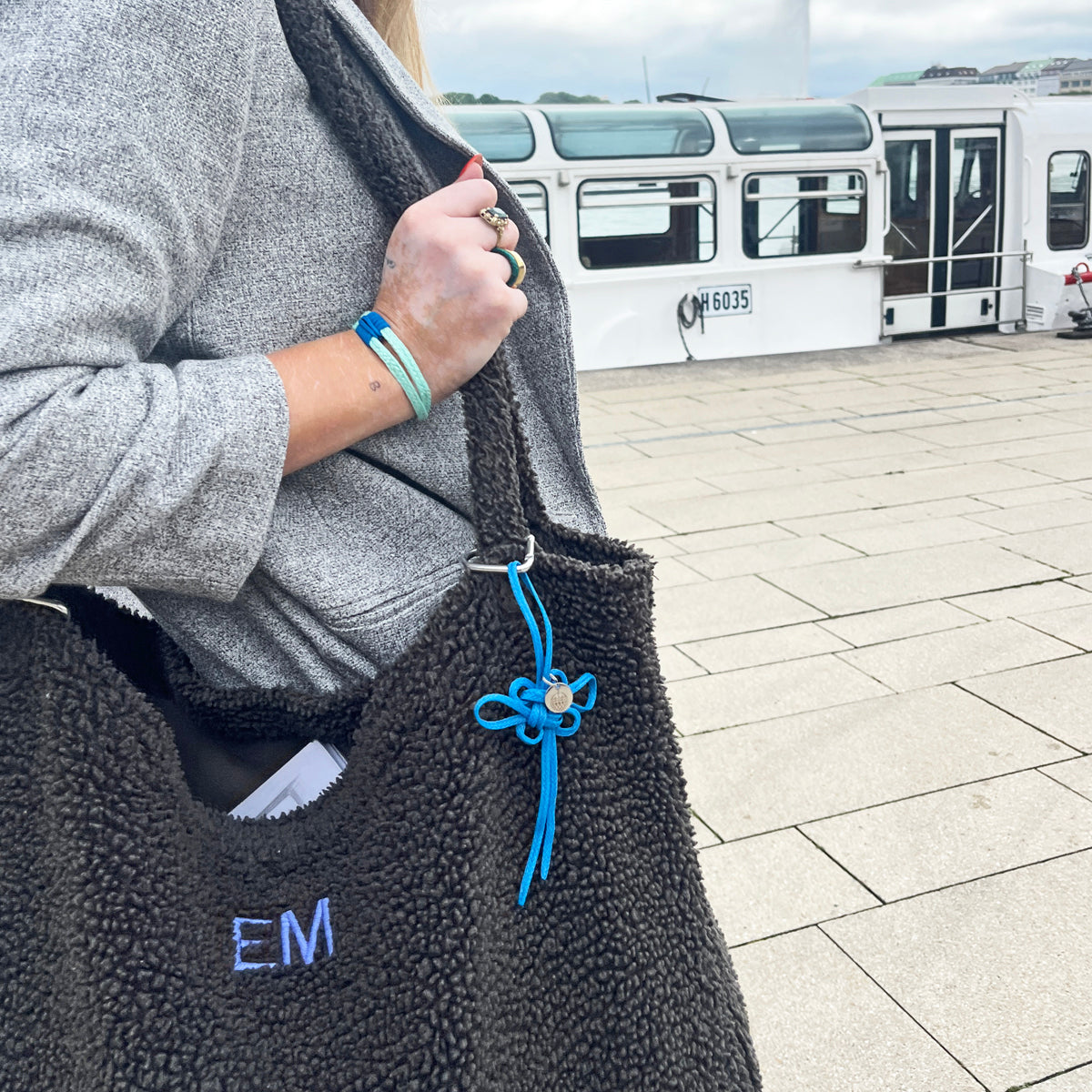 BRACENET Upcycling Taschenanhänger Flores Sea aus blauen alten Fischernetzen mit einem silber Edelstahlanhänger mit BRACENET Logo und einem silber Edelstahlclip in Herzform hängt am Henkel einer grauen Tasche. Die Tasche wird von einer Person getragen. Im Hintergrund sieht man ein Boot. 