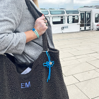 BRACENET Upcycling Taschenanhänger Flores Sea aus blauen alten Fischernetzen mit einem silber Edelstahlanhänger mit BRACENET Logo und einem silber Edelstahlclip in Herzform hängt am Henkel einer grauen Tasche. Die Tasche wird von einer Person getragen. Im Hintergrund sieht man ein Boot. 