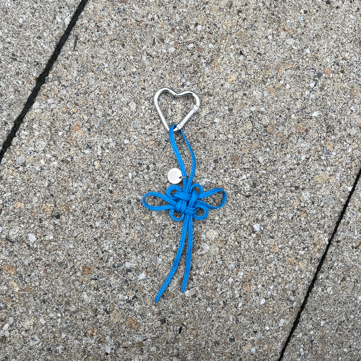 BRACENET Upcycling Taschenanhänger Flores Sea aus blauen alten Fischernetzen mit einem silber Edelstahlanhänger mit BRACENET Logo und einem silber Edelstahlclip in Herzform liegt auf Steinboden. 