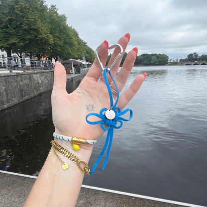 Person hält BRACENET Upcycling Taschenanhänger Flores Sea aus blauen alten Fischernetzen mit einem silber Edelstahlanhänger mit BRACENET Logo und einem silber Edelstahlclip in Herzform an einem Finger aufgehangen in der Hand. Im Hintergrund sieht man Wasser.