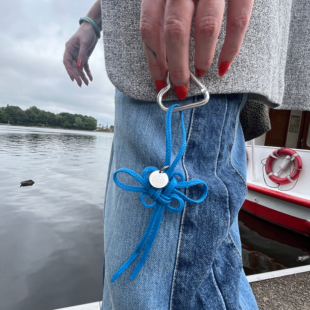 Person hält BRACENET Upcycling Taschenanhänger Flores Sea aus blauen alten Fischernetzen mit einem silber Edelstahlanhänger mit BRACENET Logo und einem silber Edelstahlclip in Herzform in der Hand und geht vor dem Wasser. 