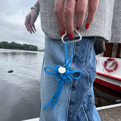 Person hält BRACENET Upcycling Taschenanhänger Flores Sea aus blauen alten Fischernetzen mit einem silber Edelstahlanhänger mit BRACENET Logo und einem silber Edelstahlclip in Herzform in der Hand und geht vor dem Wasser. 