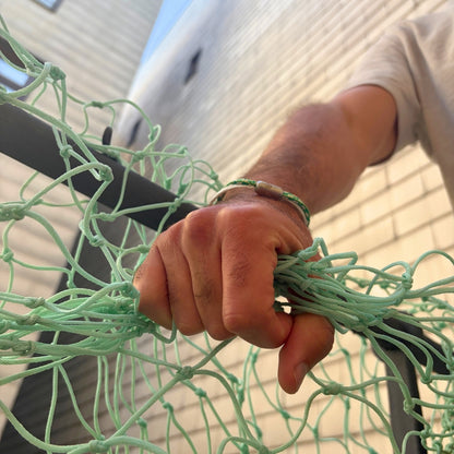 Person trägt das September Bracenet Laguna Verde aus einem Strang Fischernetz in Weiß und einem Strang Fischernetz in Grün, verbunden mit einem Knoten und einem Magnetverschluss in Silber am Handgelenk und hält Fischernetz in der Hand. 