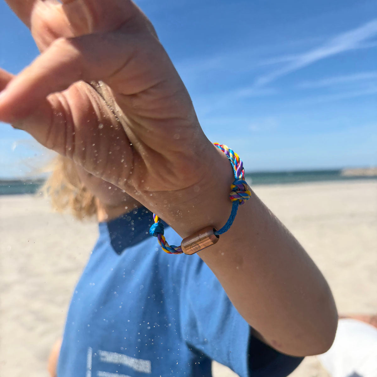 Kind ist am Strand und trägt das Kids' Bracenet Lübecker Bucht aus einem Strang blauem Fischernetz und einem Strang buntem Fischernetz, größenverstellbar mit Magnetverschluss in Roségold.  