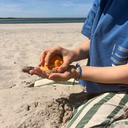 Kind trägt das Kids' Bracenet Lübecker Bucht aus einem Strang blauem Fischernetz und einem Strang buntem Fischernetz, größenverstellbar mit Magnetverschluss in Roségold am Handgelenk und hat ein Sandförmchen in der Hand. 