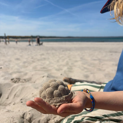 Kind trägt das Kids' Bracenet Lübecker Bucht aus einem Strang blauem Fischernetz und einem Strang buntem Fischernetz, größenverstellbar mit Magnetverschluss in Roségold am Handgelenk und hat eine Sandfigur in der Hand. 