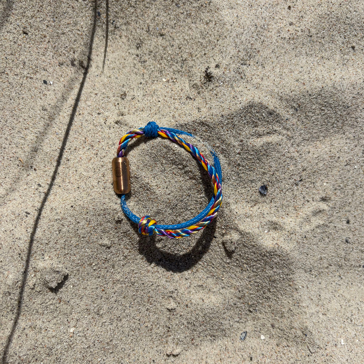 Das Kids' Bracenet Lübecker Bucht aus einem Strang blauem Fischernetz und einem Strang buntem Fischernetz, größenverstellbar mit Magnetverschluss in Roségold liegt im Sand. 