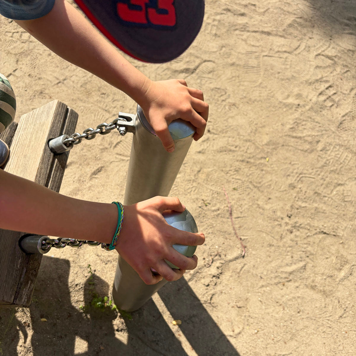 Kind trägt das Kids' Bracenet Kieler Bucht aus einem Strang türkisen Fischernetz und einem Strang buntem Fischernetz, größenverstellbar mit Magnetverschluss in Silber am Handgelenk und ist auf einem Spielplatz. 
